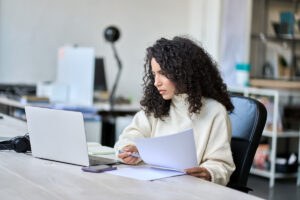 A woman reviewing her taxes on a laptop and paperwork at a desk.